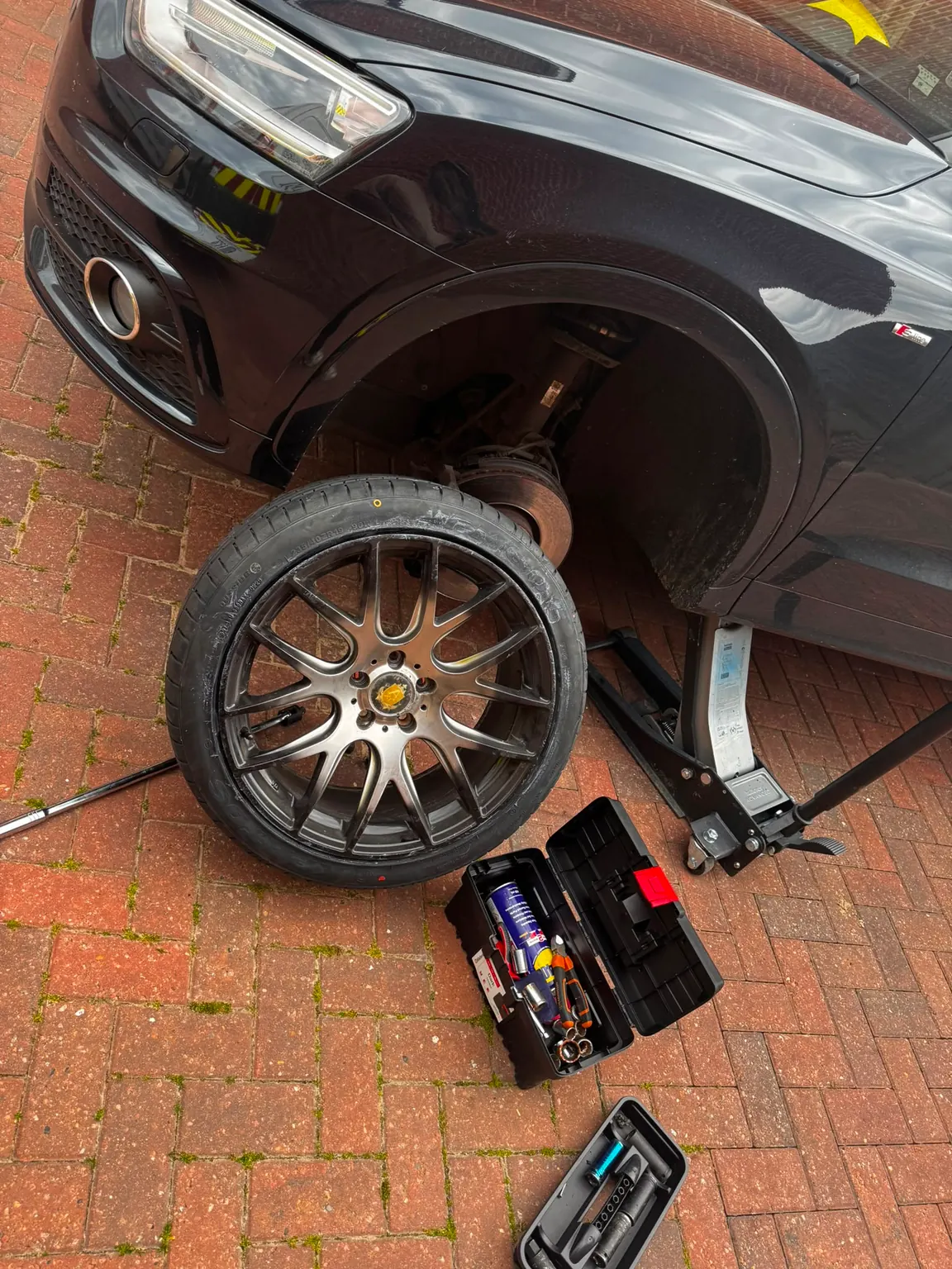 A large alloy wheel resting on a block-paved driveway beside the black car it came off, which is raised on a jack.