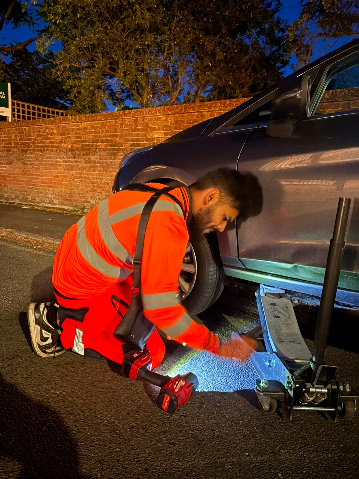 A Fly Tyres fitter in high-visibility orange positioning a trolley jack under a car on an unlit residential street at night.