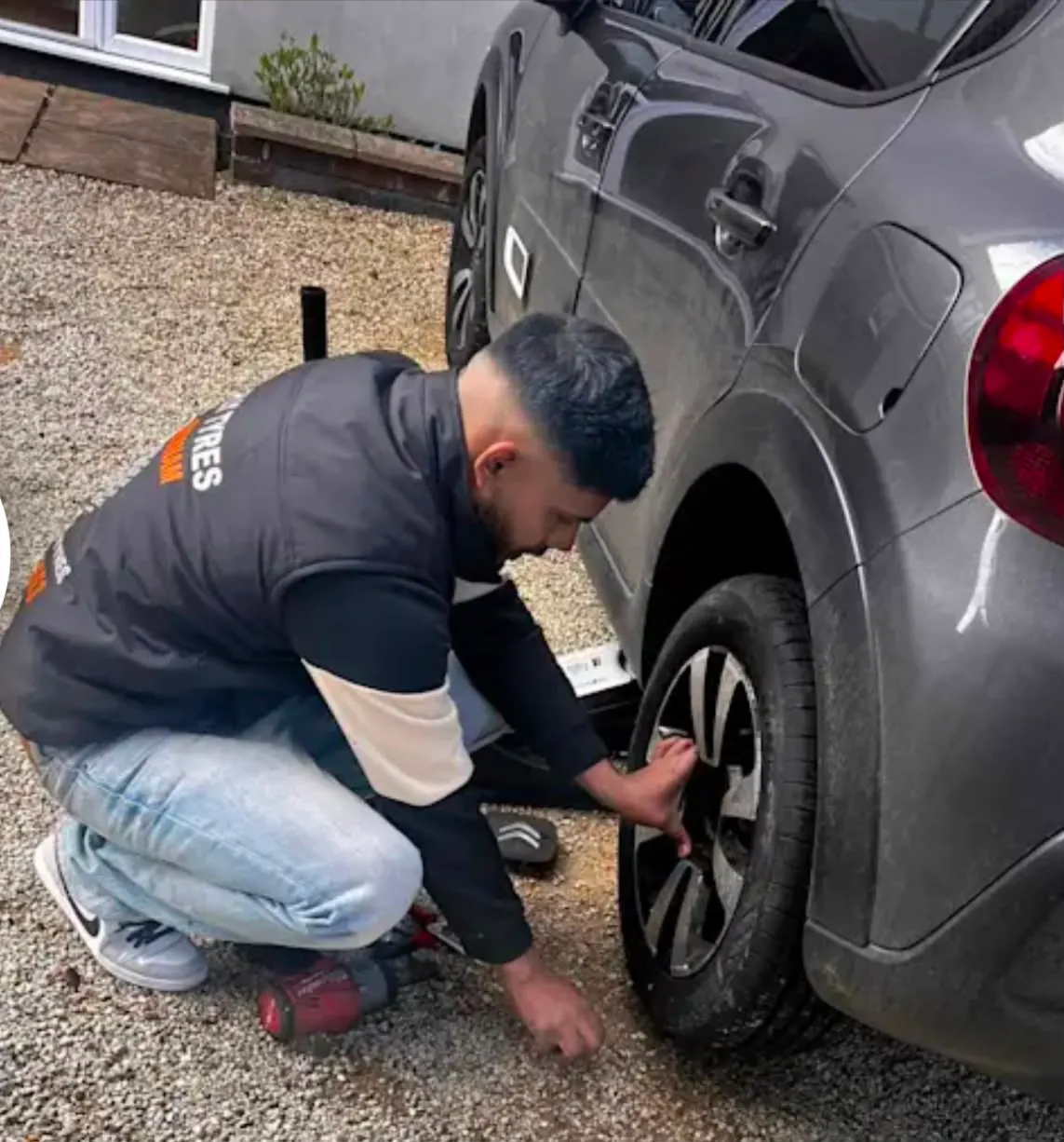 A Fly Tyres fitter crouched at the rear wheel of a grey car on a gravel driveway, loosening the wheel nuts.