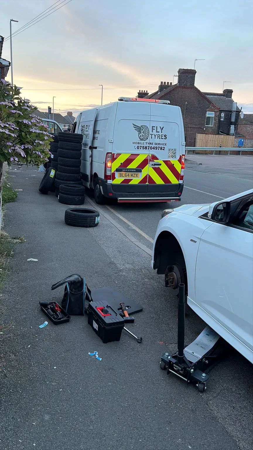 A car jacked up at the kerb with its wheel removed, tools laid out on the pavement and the Fly Tyres van parked ahead of it.