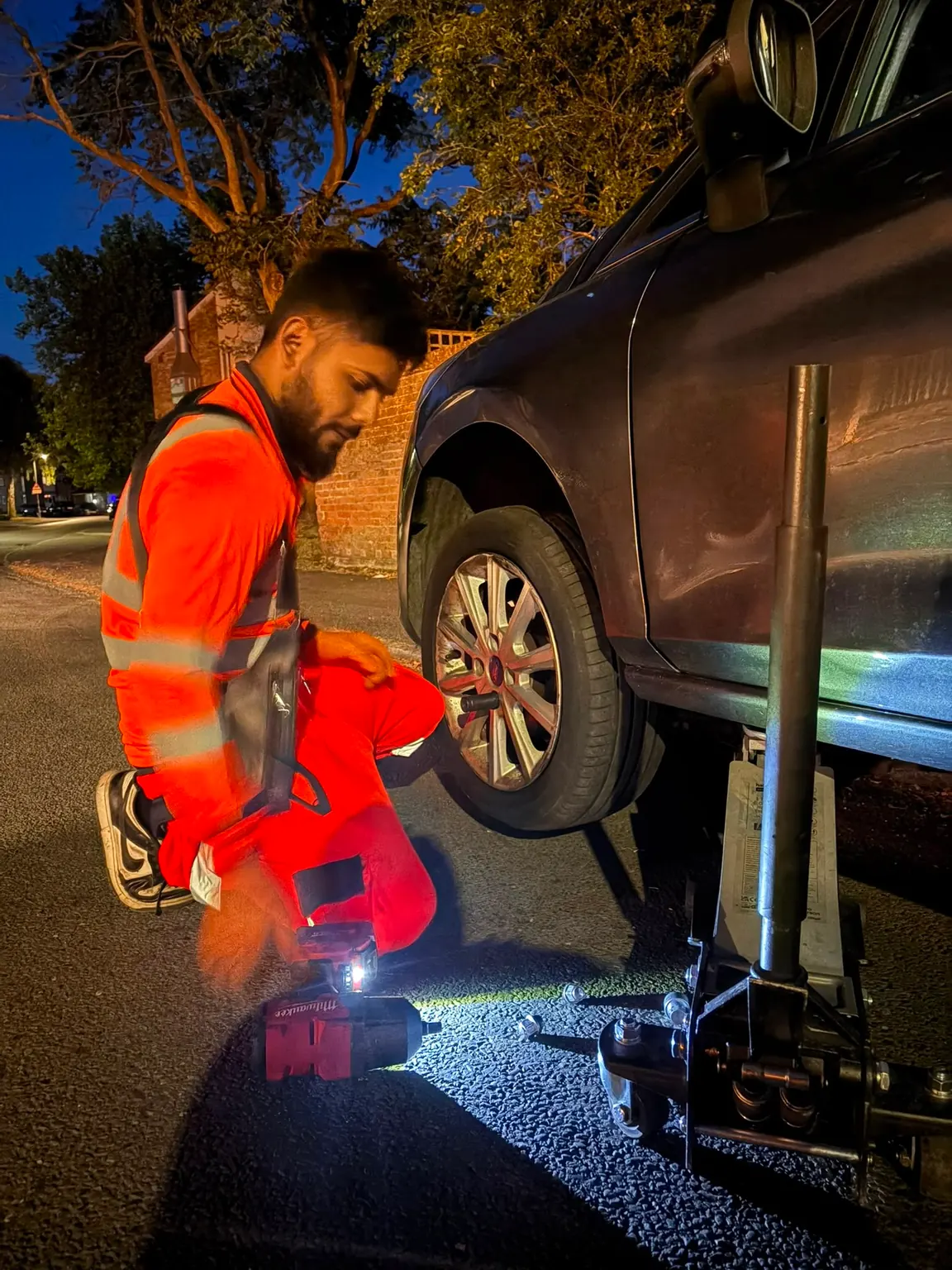 A Fly Tyres fitter in a high-visibility orange suit kneeling beside a car wheel at night, working by the light of a hand lamp.