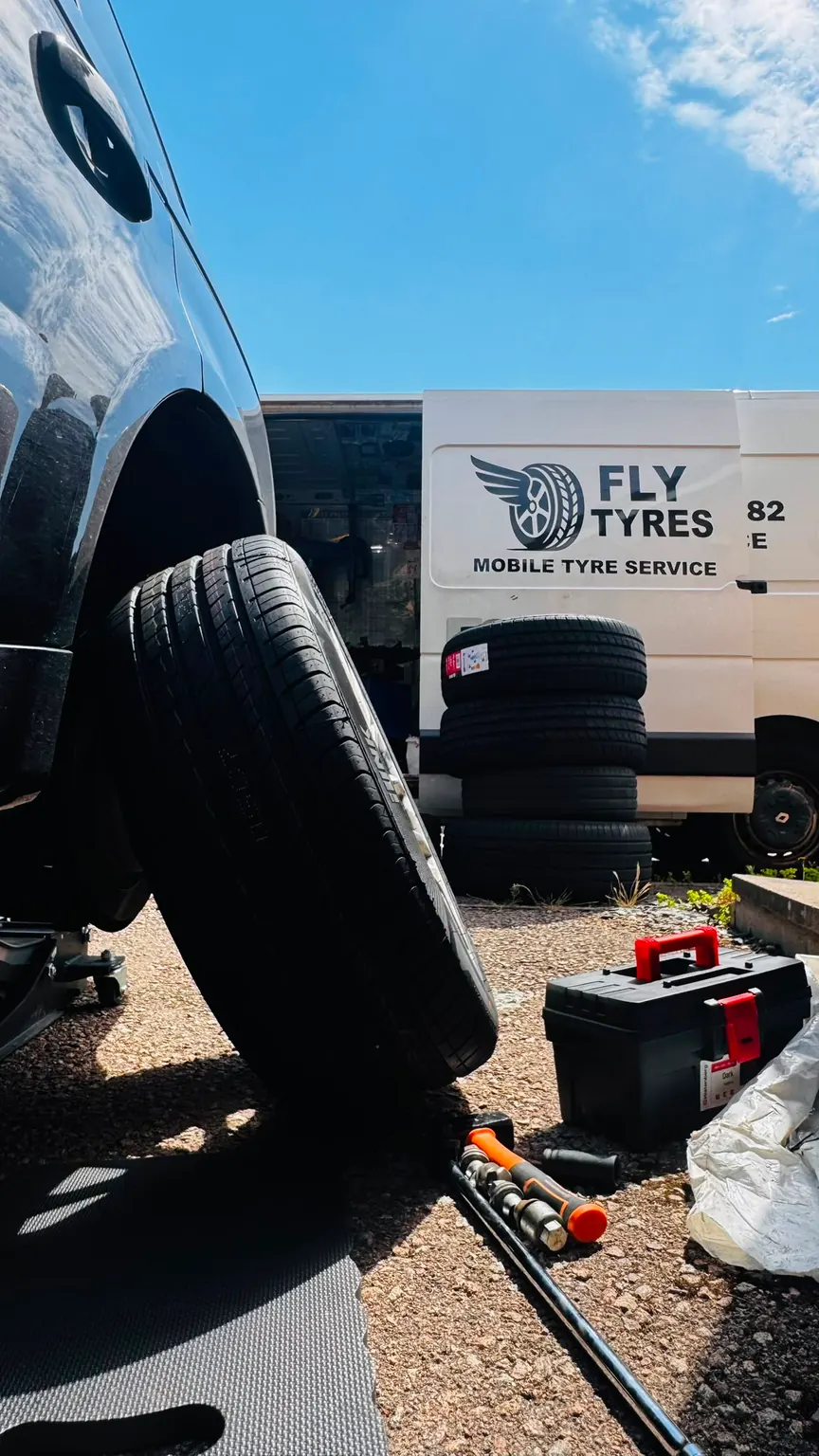 A new tyre stood upright against a car with its wheel removed, the Fly Tyres van and toolbox behind it in bright sunshine.