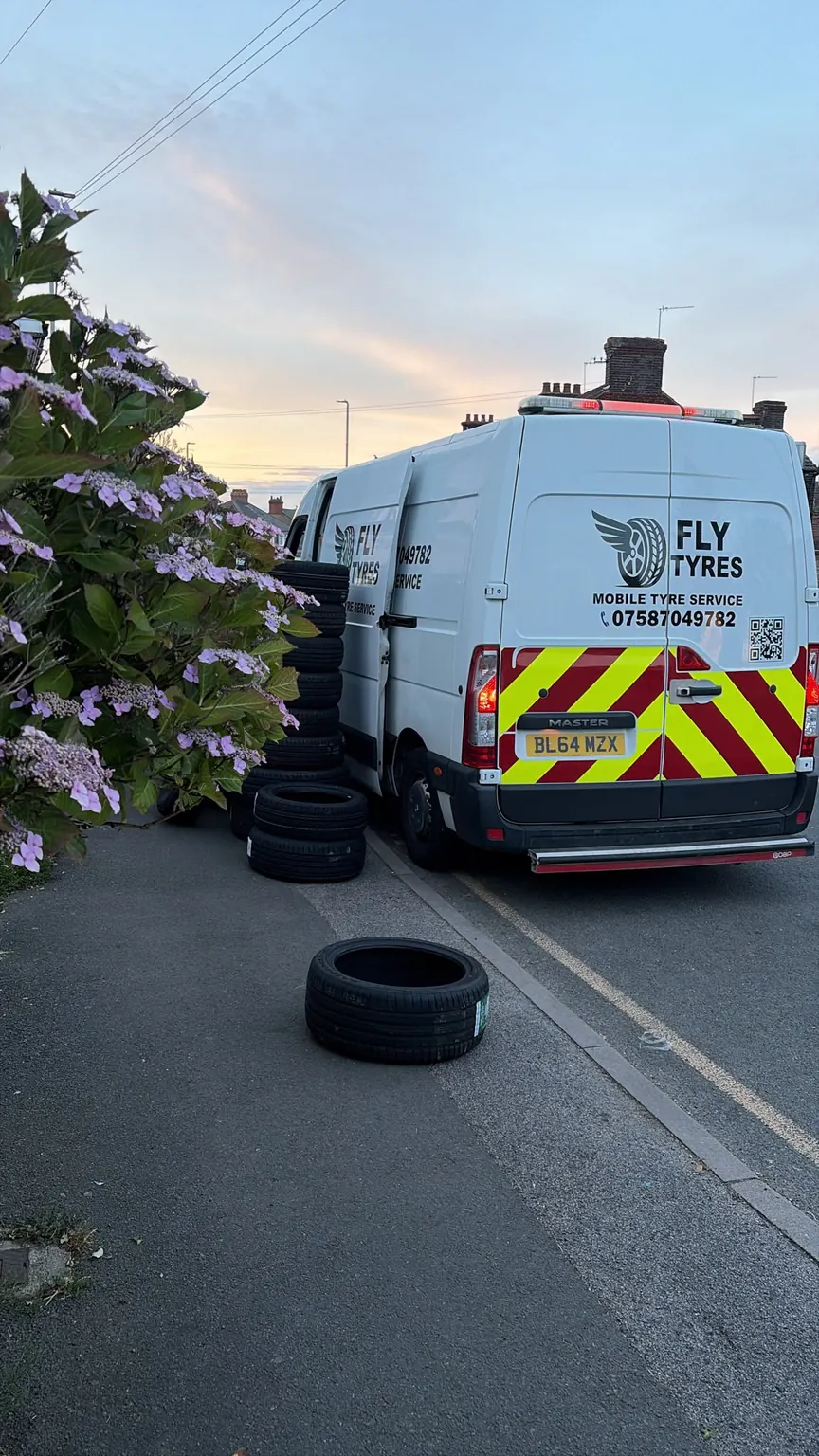 The rear of the Fly Tyres van parked on a residential road at dawn, with replacement tyres stood on the kerb behind it.